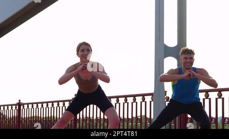Fröhlicher Mann und Frau in Sportbekleidung trainieren zusammen im Freien und machen seitliche Ausfallschritte. Kaukasische Sportler und Sportlerpaare, die Sport treiben Stockfoto