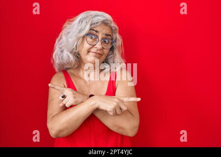 Eine Frau mittleren Alters mit grauen Haaren, die auf rotem Hintergrund steht und mit den Fingern auf beide Seiten zeigt, unterschiedliche Richtungen stimmen nicht überein Stockfoto