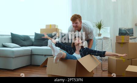 Ein junges Paar, das sich in einer neu gekauften Wohnung amüsiert. Glücklicher Mann und Frau, die am Umzugstag spielen. Ein hübscher Mann, der mit seiner Freundin Pappschachtel schiebt Stockfoto