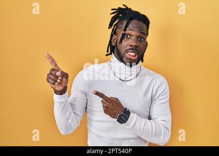 Ein afrikanischer Mann mit Dreadlocks, der einen Rollkragenpullover auf gelbem Hintergrund trägt und zur Seite zeigt, besorgt und nervös mit beiden Händen, besorgt und surpri Stockfoto