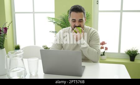 Junger latino-Mann, der Apfel isst und zu Hause ein Notebook benutzt Stockfoto