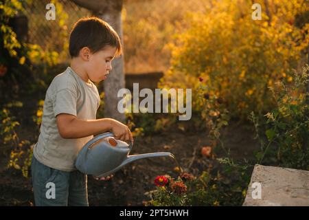 Ein kleiner Junge mit einer Gießkanne und Blumen aus dem Garten an sonnigen Tagen. Süße kleine Kindergärtnerei, im Sommer im Garten. Familienheim Freizeit Stockfoto