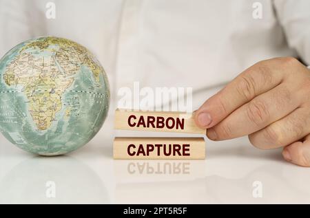 Auf einer weißen reflektierenden Oberfläche befindet sich ein Globus in den Händen von Holzblöcken mit der Aufschrift „Carbon Capture“. Das Konzept von Industrie und Umwelt Stockfoto