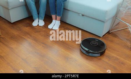 Moderne Staubsauger-Reinigung zu Hause. Schließen. Sauberer Staubsauger Parkett im Zimmer. Das Paar hebt die Beine, während es auf der Couch sitzt. Großer Blickwinkel Stockfoto