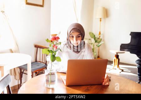 Eine hübsche junge Frau aus dem Nahen Osten, die zu Hause Hijab mit einem Laptop trägt. Ein muslimisches Mädchen bereitet sich auf die Aufnahmeprüfung vor. Heimunterricht und Online-Studium-Konzept Stockfoto