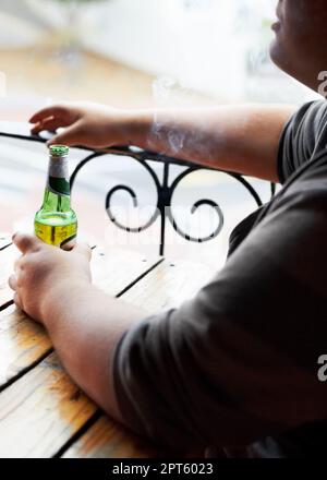 Leben genießen, kleine Freuden. Ein abgeschnittenes Bild eines jungen Mannes, der raucht, während er mit einem Bier vor sich sitzt Stockfoto