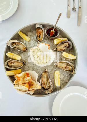 Leckere frische Austern mit Zitrone auf Eis, Käse mit Gewürzen und leckeres Brot. Serviert mit Messern, Gabel, Löffel und Teller. Weiß Stockfoto