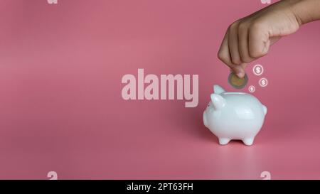 Eine menschliche Hand steckte eine Münze in ein Sparschwein auf einem rosa Hintergrund. Einsparungs- und Investitionskonzepte. Stockfoto