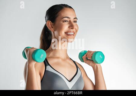 Lasst den Gewinn beginnen. Studioaufnahme einer sportlichen jungen Frau, die mit Kurzhanteln vor einem grauen Hintergrund trainiert Stockfoto