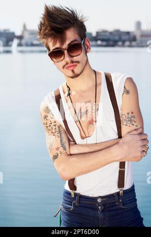 Das Künstlerleben zu leben. Ein Hipster in Hosenträgern und mit Tattoos auf den Armen und einer Sonnenbrille, die draußen steht Stockfoto