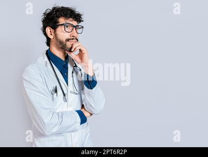 Junger Arzt, der isoliert denkt und aufblickt. Ein pensiver Arzt, der isoliert aufblickt, ein Doktor, der mit der Hand am Kinn denkt Stockfoto
