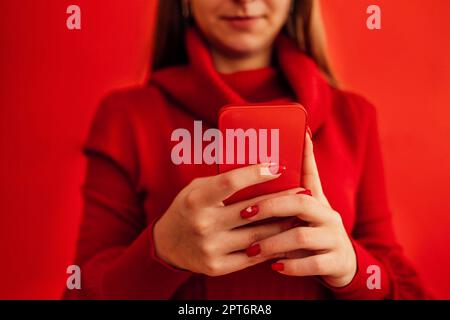 Blondes, lächelndes Teenager-Mädchen in einem roten Pullover hält ein rotes Telefon in der Hand. Nahaufnahme einer wunderschönen, leuchtend roten Maniküre. Speicherplatz kopieren. Isoliert auf einem Stockfoto