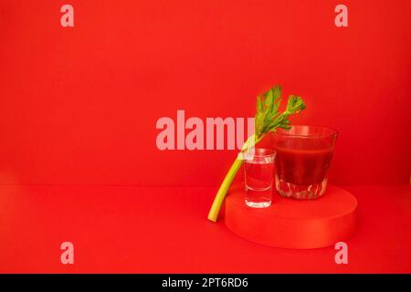 Ein Glas roter Cocktail mit Grün auf dem Podium. Stilvolles alkoholisches Tomatengetränk Bloody Mary auf rotem Hintergrund. Köstliches Getränkekonzept. Verstanden Stockfoto