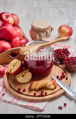 Stillleben mit frischen Preiselbeeren, roten Äpfeln mit Messer auf Holzbrett und Glasbecher mit hausgemachter Marmelade auf hellem Hintergrund. Herbsternte, Stockfoto