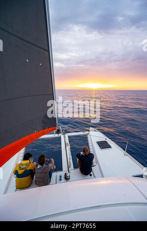 Drei junge Männer sitzen auf dem Katamaran und schauen auf den Sonnenuntergang, Rhodos, Dodekanese, Griechenland Stockfoto