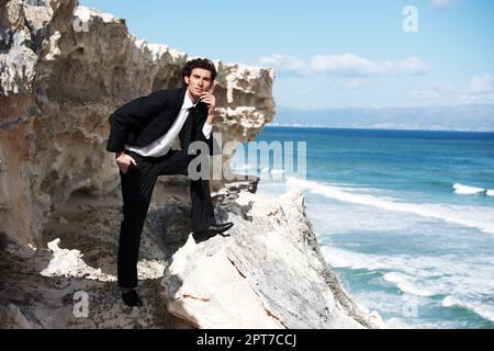 Was für eine schöne Aussicht... ein gutaussehender Geschäftsmann, der auf einem Felsen mit Blick auf das Meer für die Kamera posiert Stockfoto