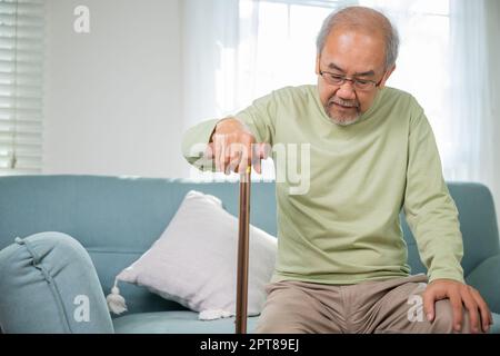 Asiatischer alter Mann mit einer Brille, die mit Gehstock vom Sofa aus aufsteht, ältere Menschen mit Knieschmerzen, die den Griff des Rohrs halten, Senior DIS Stockfoto