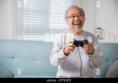 Asiatische ältere alte Mann genießen halten Joystick spielen Videospiel zu Hause im Wohnzimmer, reifen Mann Hände mit Game-Controller, Funny auf Ruhestand Stockfoto