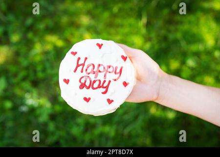 Bento-Kuchen mit der Aufschrift Happy Day und roten Herzen hält ein Mädchen einen Kuchen in der Hand auf einem Hintergrund aus grünem Gras. Sonniger Tag. Selektiver Fokus Stockfoto