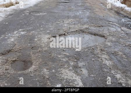 Beschädigte Asphaltstraße mit Schlaglöchern durch Gefrier- und Auftauzyklen im Winter. Schlechte Straße Stockfoto