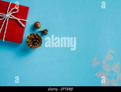 Weihnachtshintergrund: Rote Geschenkbox mit weißem und silbernem Kordelband, Nadelzapfen und Eichel auf blauem Papierhintergrund. Flaches Layout mit Kopie Stockfoto