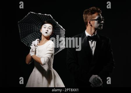 Portrait von Paar mime mit Sonnenschirm auf schwarzen Hintergrund. Mann im Smoking und Gläser und Frau im weißen Kleid. Beziehung Stockfoto