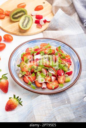 Vegetarisches Obst und Gemüse Salat aus Erdbeere, Kiwi, Tomaten, mikrogrünen Sprossen auf weißem Beton Hintergrund und Leinentextilien. Seitenansicht, Kl Stockfoto