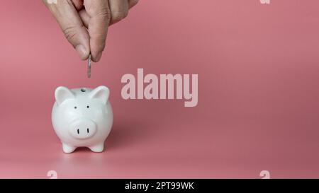 Eine menschliche Hand lässt eine Münze in ein weißes Sparschwein fallen. Auf einem rosa Hintergrund. Einsparungs- und Investitionskonzepte. Stockfoto