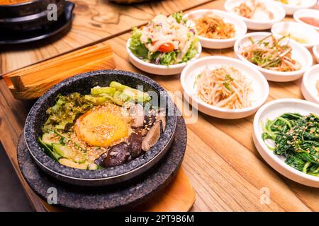 Bibimbap in einer schwarzen, beheizten Steinschale, koreanisches traditionelles Gericht - Bibimbap gemischter Reis mit Gemüse umfassen Rindfleisch und gebratenes Ei und Gurken auf Tisch gesetzt Stockfoto