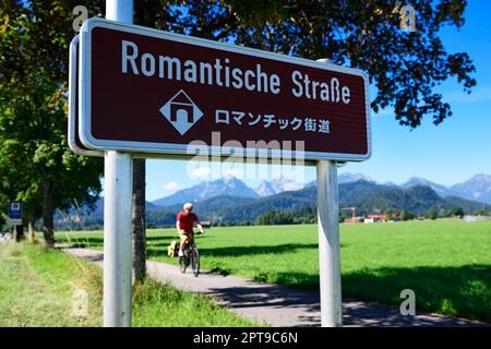 Radfahrer vor dem Schild Romantische Straße mit Übersetzung in Japanisch, Schwangau, Oberbayern, Bayern, Deutschland Stockfoto