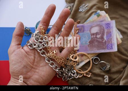 Marodeuter reichen mit einem Haufen gestohlenem Schmuck und Geld auf Militäruniform-Stoff und russischer Flagge. Plünderung durch russische Soldaten in der Ukraine Stockfoto