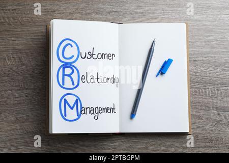 Notizbuch mit Abkürzung CRM (Customer Relationship Management) und Stift auf Holzhintergrund, Draufsicht Stockfoto