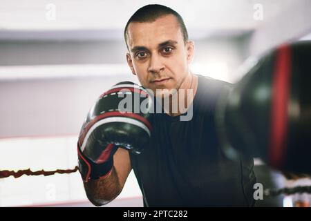 Mit den Schlägen. Porträt eines selbstbewussten jungen Boxers, der Boxhandschuhe trägt und in einem Ring einen gy mit Schlägen gegen die Kamera wirft Stockfoto