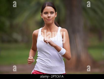 Sie sprang durch den Park zu ihrem Lieblingslied. Eine attraktive brünette Frau, die in Sportkleidung durch den Park rennt Stockfoto