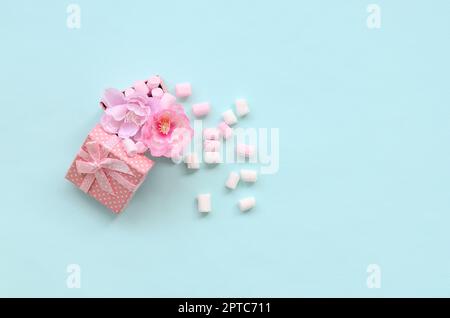 Rosa Geschenkkarton mit Blumen bestreut mit Marshmallows auf blauem Hintergrund. Metaphorische Konzept, die Verarbeitung, den Anbau von Blumen. Minimalismus flach Stockfoto