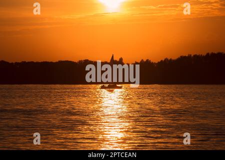 Segelboot bei Sonnenuntergang auf dem Fluss. Das Boot fährt entlang eines Lichtwegs von der untergehenden Sonne. Stockfoto