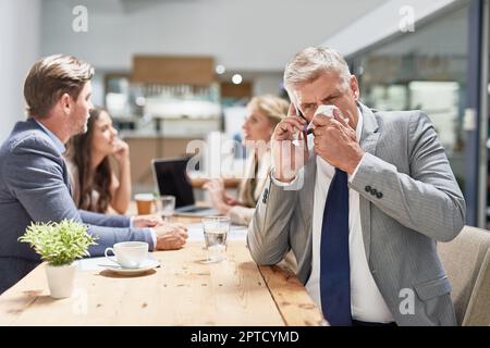 Warten Sie nicht, bis es Ihnen schlechter geht, bevor Sie zum Arzt gehen. Ein Geschäftsmann, der sich die Nase putzt, während er in einem Büro mit einem Handy spricht Stockfoto