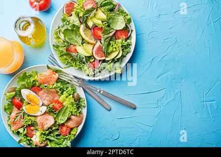 Frische grüne Salatsalate mit geräuchertem Lachs, Kirschtomaten, Ei und Avocado auf blauem Tischhintergrund Stockfoto