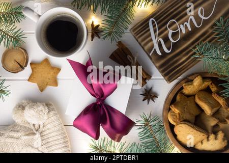 Startseite Schriftzug verbranntes Holzschild, Lebkuchenkeks, Christbaumzweige, Tasse Kaffee, Geschenk, Zimtstangen auf weißer Oberfläche. Flach liegend Stockfoto