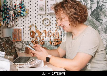 Lächelnde, konzentrierte Geschäftsfrau, die Smartphone spricht, am Laptop arbeitet, Ordnung nimmt. Babyzubehör, Silikon-Öko-Rasseln, Beruhigungssauger, Stockfoto
