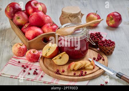 Stillleben mit frischen Preiselbeeren, roten Äpfeln mit Messer auf Holzbrett und Glasbecher mit hausgemachter Marmelade auf hellem Hintergrund. Herbsternte, Stockfoto