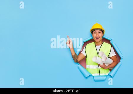 Der junge asiatische Ingenieur, isoliert über blauem Hintergrund, trägt einen gelben Schutzhelm und hält Papierrollen in der Hand, zeigt mit dem Finger auf freien Spachtel Stockfoto