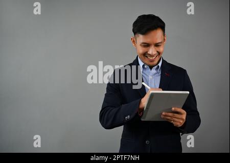 Professioneller und erfolgreicher asiatischer Millennials-Geschäftsmann in formellen Business-Anzügen, der sein digitales Tablet auf einem isolierten grauen Hintergrund verwendet Stockfoto