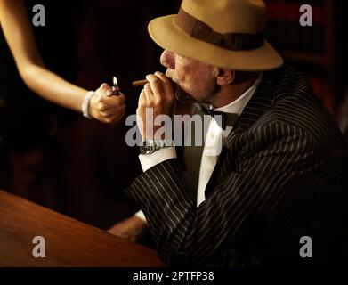 Er hat Macht und Einfluss über andere. Der alte Mafia-Boss trägt einen Hut und sieht ernst aus, während eine Frau ihm eine Zigarette anzündet Stockfoto