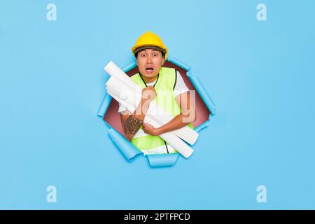 Ein junger ängstlicher Mann, der isoliert vor blauem Hintergrund postet und einen gelben Schutzhelm trägt, ein paar Papierrollen in der Hand hält, einen Kopierraum, ein hochwertiges Foto. Stockfoto