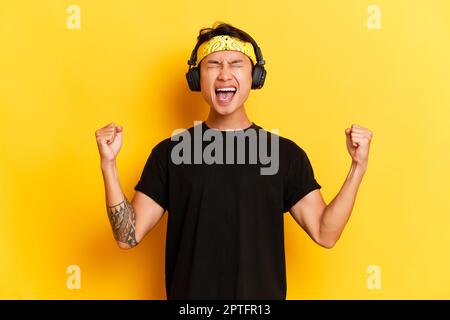 Porträt eines jungen, glücklichen Mannes in schwarzem T-Shirt, der auf gelbem Studiohintergrund posiert. Er trägt Kopfhörer und ein gelbes Stirnband, weint fröhlich und hat Urlaub Stockfoto
