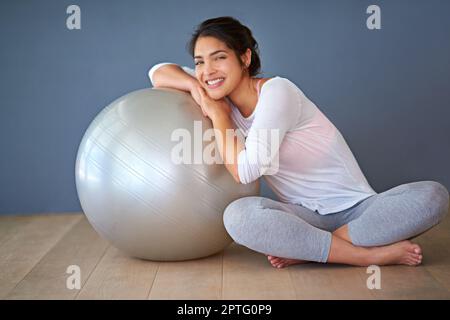 Liebe dich genug, um ein gesundes Leben zu führen. Gesamtbild einer sportlichen jungen Frau, die sich vor grauem Hintergrund auf einen Pilates-Ball lehnt Stockfoto