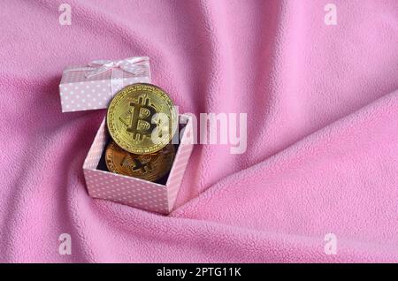 Die goldenen bitcoin liegt in einem kleinen rosa Geschenkkarton mit einer kleinen Verbeugung auf einer Decke aus weichem, flauschigen Rosa fleece Stoff mit einer großen Anzahl von Stockfoto