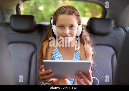 Sie sah sich Filme auf dem Rücksitz an. Ein junges Mädchen, das auf dem Rücksitz sitzt, Kopfhörer trägt und ein digitales Tablet benutzt Stockfoto