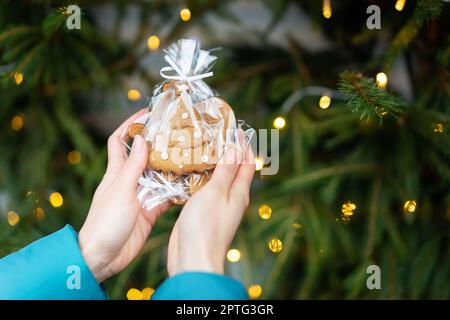 Eine Frau hält in ihren Händen Lebkuchen in Form eines Weihnachtsbaums auf dem Hintergrund eines grünen Weihnachtsbaums im Freien. Das Konzept des Winterhols Stockfoto